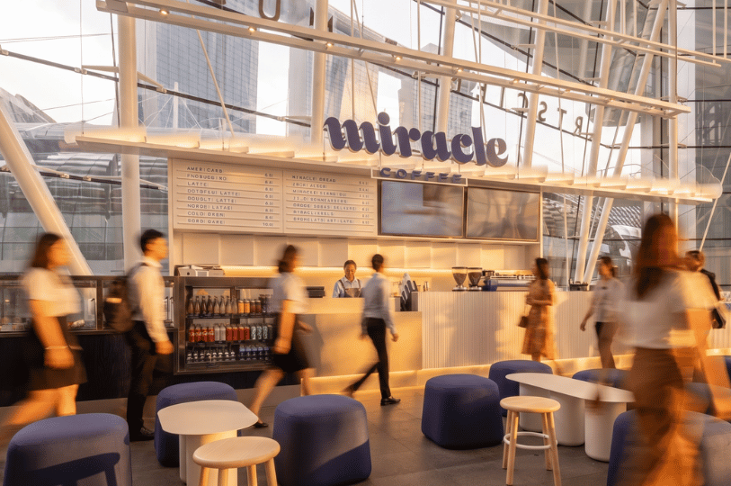 Pedestrians blur in motion as they move past Miracle Coffee, a bright, open-concept cafe with blue seating nestled under a striking modern glass architectural structure.
