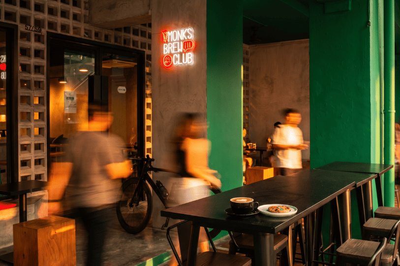 A focused shot of a coffee cup and cookie on a dark table outside Monks Brew Club, while a cyclist and pedestrians rush past its green pillars and glowing neon sign in a blur.