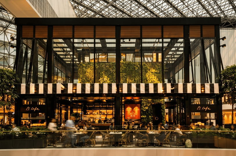 Diners are seated inside a grand, two-story glass-enclosed PS.Cafe located within a bright mall atrium, contrasted by the fast-paced, blurred motion of shoppers passing by outside.