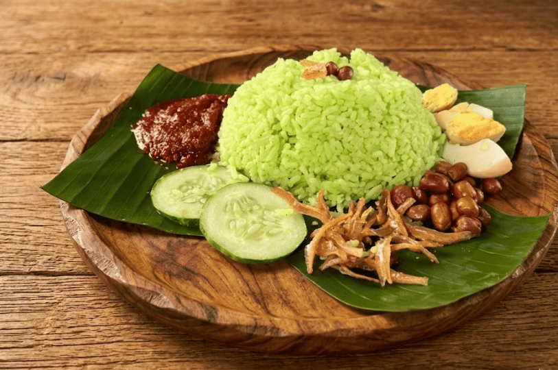 A vibrant green pandan-infused Nasi Lemak rice set served on a wooden plate, accompanied by traditional sides of sambal, sliced cucumbers, fried anchovies, peanuts, and a hard-boiled egg.