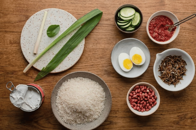 A top-down flat lay of raw Nasi Lemak ingredients including a bowl of long-grain rice, a can of coconut milk, pandan leaves, lemongrass, fresh cucumber slices, sambal, fried ikan bilis, peanuts, and boiled eggs.