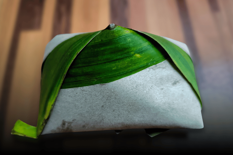 A classic "Nasi Lemak Bungkus" wrapped tightly in traditional brown paper and a signature green banana leaf, forming a small pyramid shape ready for takeaway.