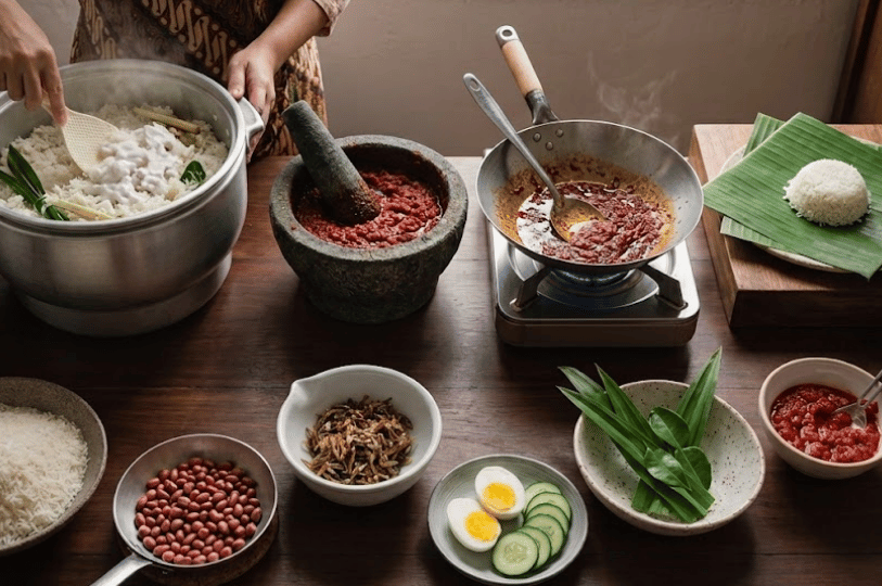 A wide shot of a kitchen prep station showing the labor-intensive process of making Nasi Lemak, including a large pot of coconut rice, a mortar and pestle for sambal, and various bowls of prepared condiments.