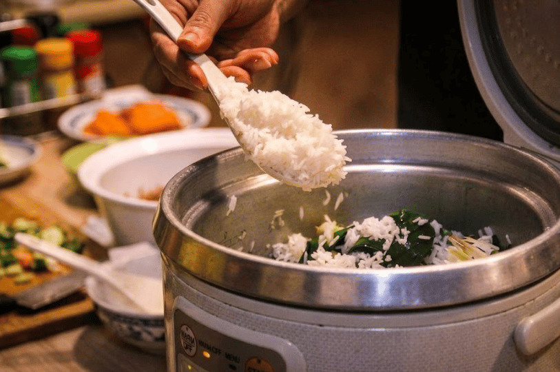 A close-up of a cook fluffing fragrant white coconut rice in a commercial rice cooker, with pandan leaves visible inside to illustrate the traditional steaming method.
