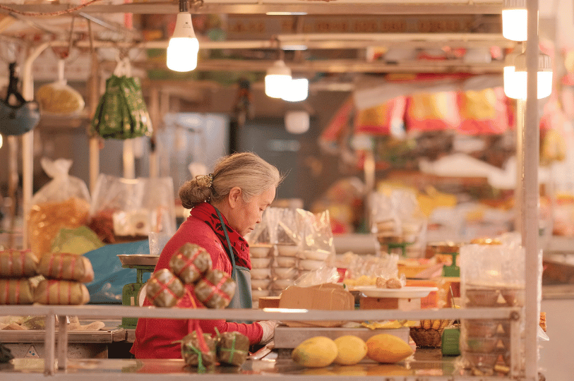 An older woman with gray hair, wearing a red shirt, works diligently at her brightly lit, well-stocked market stall. The counter displays traditional wrapped food bundles, fresh mangoes, and a green weighing scale, illustrating an active and thriving local business.